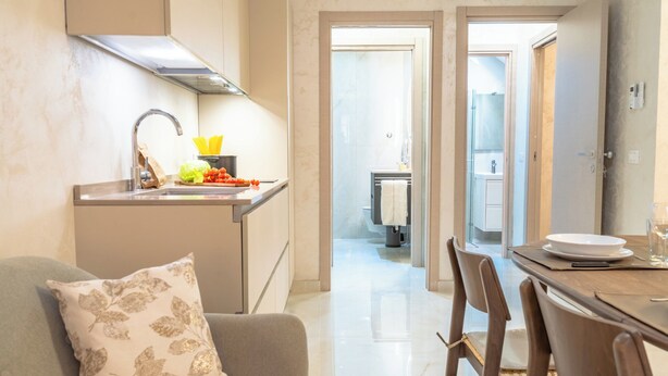 Cozinha moderna com fogão embutido, pia e bancada com legumes frescos. Há uma área de jantar com mesa e cadeiras, e uma sala de estar com sofá e almofada decorativa.