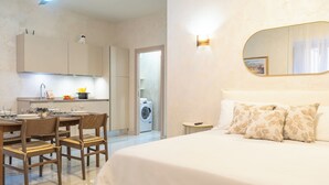 Quarto de hotel moderno com uma cama grande, área de refeições com mesa e cadeiras, cozinha com máquina de lavar e banheiro.