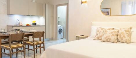 A modern hotel room with a large bed, a dining area with a table and chairs, a kitchen with a washing machine, and a bathroom.