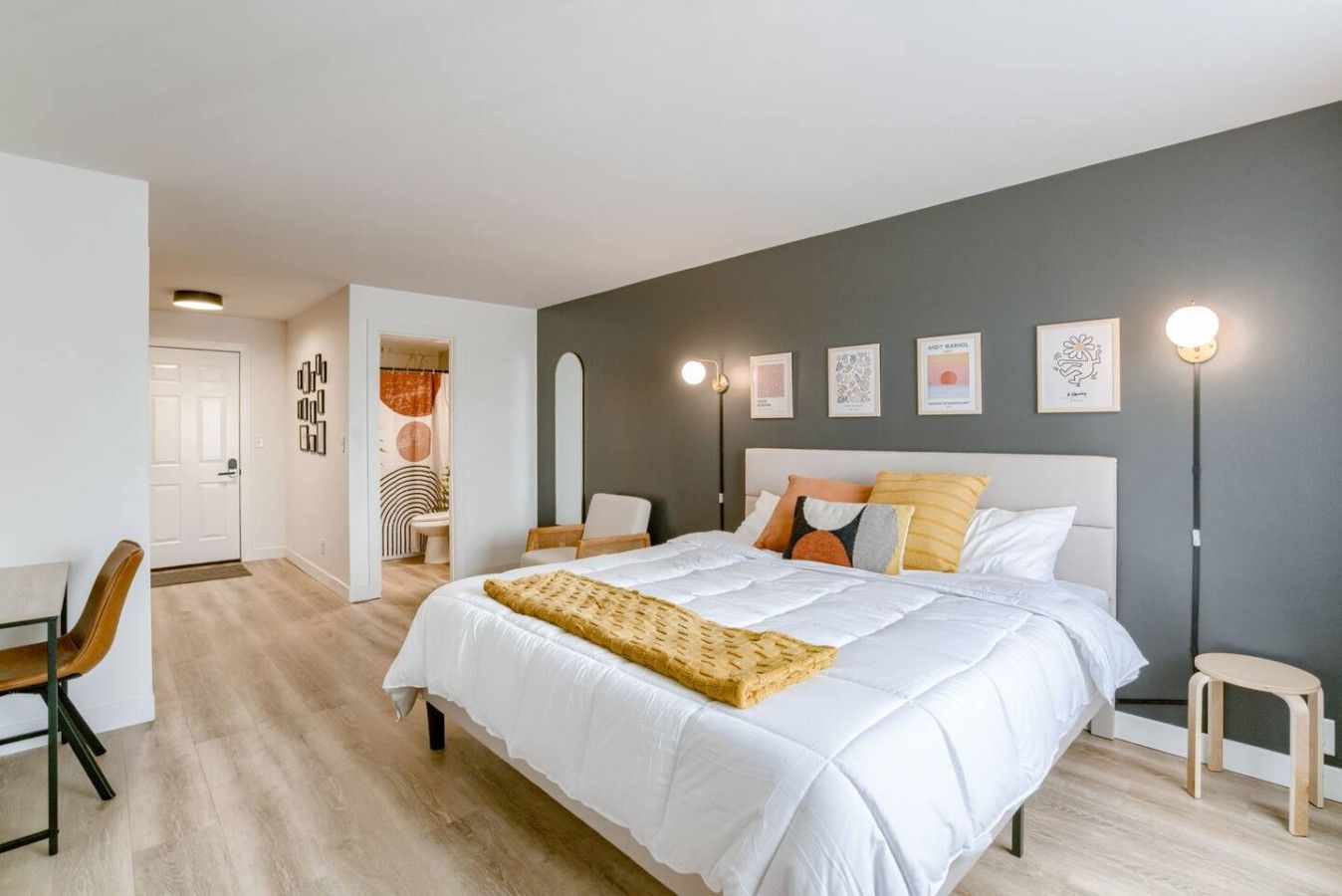 A modern bedroom with a large bed, a desk, and a chair. There are wall-mounted lights and framed pictures.