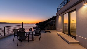Una terrazza con tavolo e sedie, una poltrona sospesa e vista sul mare al tramonto.