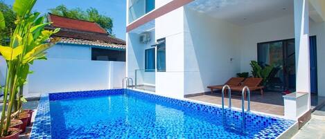 A modern pool area with a tiled blue pool, a covered seating area, and potted plants.
