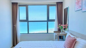 Une chambre d’hôtel avec une grande fenêtre, un lit avec une literie blanche, un oreiller rose et un vase rempli de fleurs roses posé sur une table de chevet.