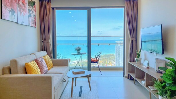 Un salon moderne avec un canapé, une table basse et un téléviseur. Un balcon avec vue sur la mer est visible à travers les portes vitrées.