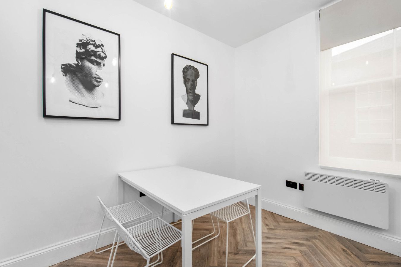 A minimalist room with a white table, two framed artworks, a radiator, and a window.