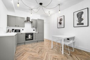 A modern kitchen with grey cabinets, a white countertop, and a herringbone wood floor. There is a white dining table with chairs and two framed artworks on the wall. - Modern 3 Bedroom Townhouse - Bedroom TVs - Wolverhampton Centre (Wolverhampton)