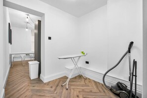 Una habitación limpia y luminosa con piso de madera en espiga, un pequeño centro de planchado y una aspiradora.