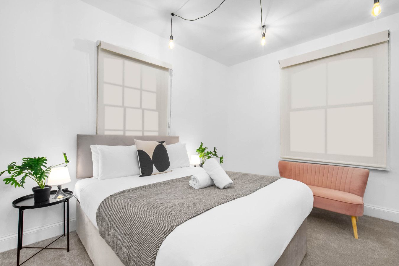A modern hotel room with a large bed, a bedside table with a lamp, a potted plant, and a pink sofa.