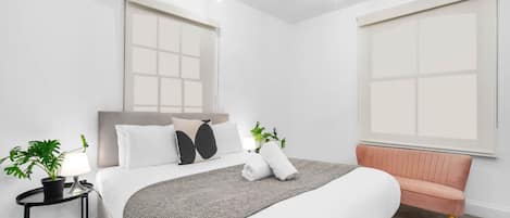 A modern hotel room with a large bed, a bedside table with a lamp, a potted plant, and a pink sofa.