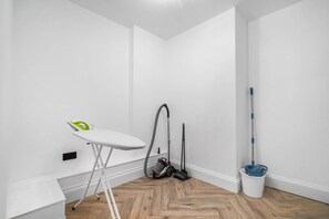 Una habitación pequeña y limpia con piso de madera, una tabla de planchar blanca, un aspirador y un trapeador.