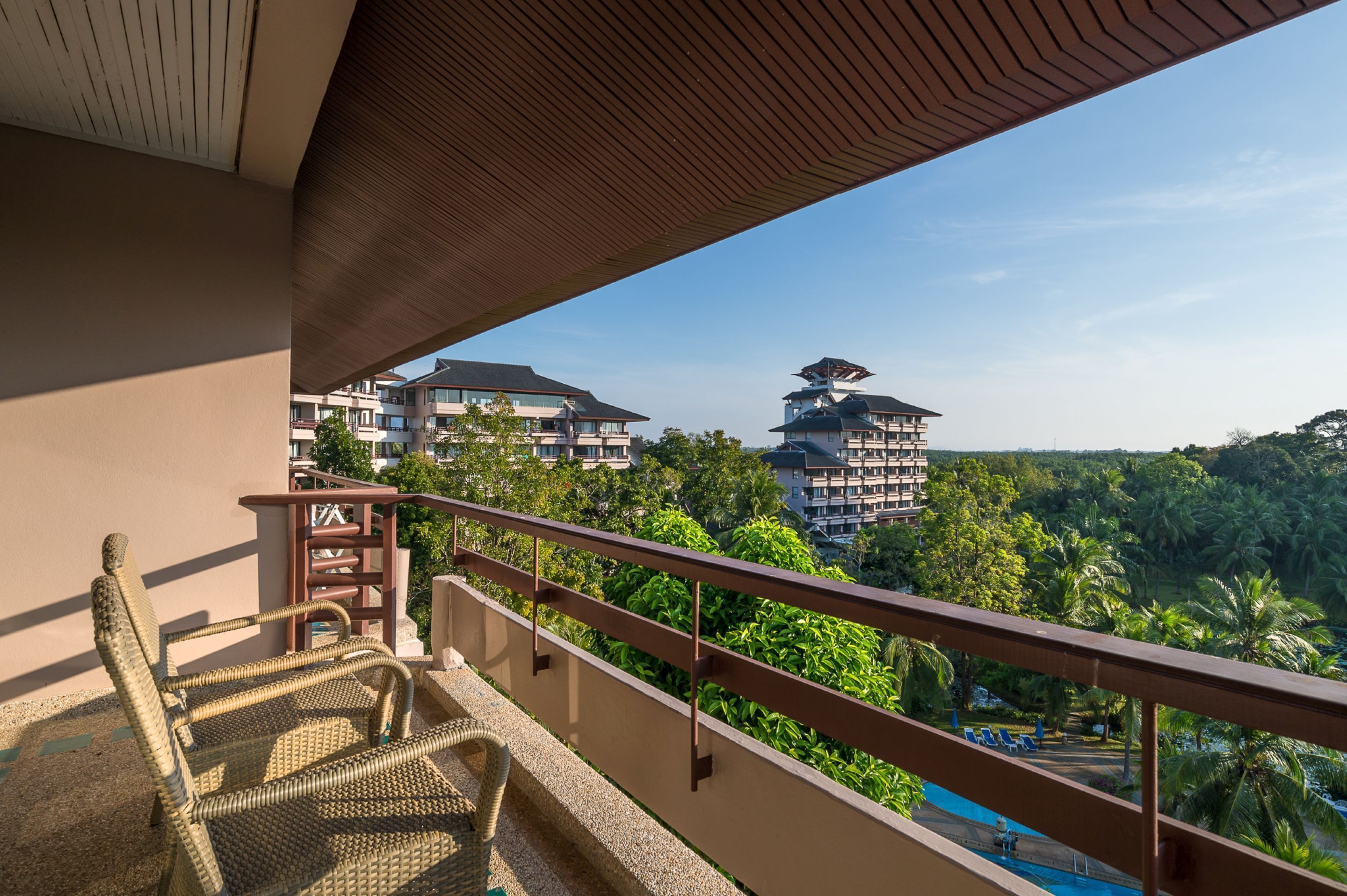 Een balkon met rieten stoelen, een houten tafel en uitzicht op een resort met meerdere gebouwen en weelderige begroeiing.
