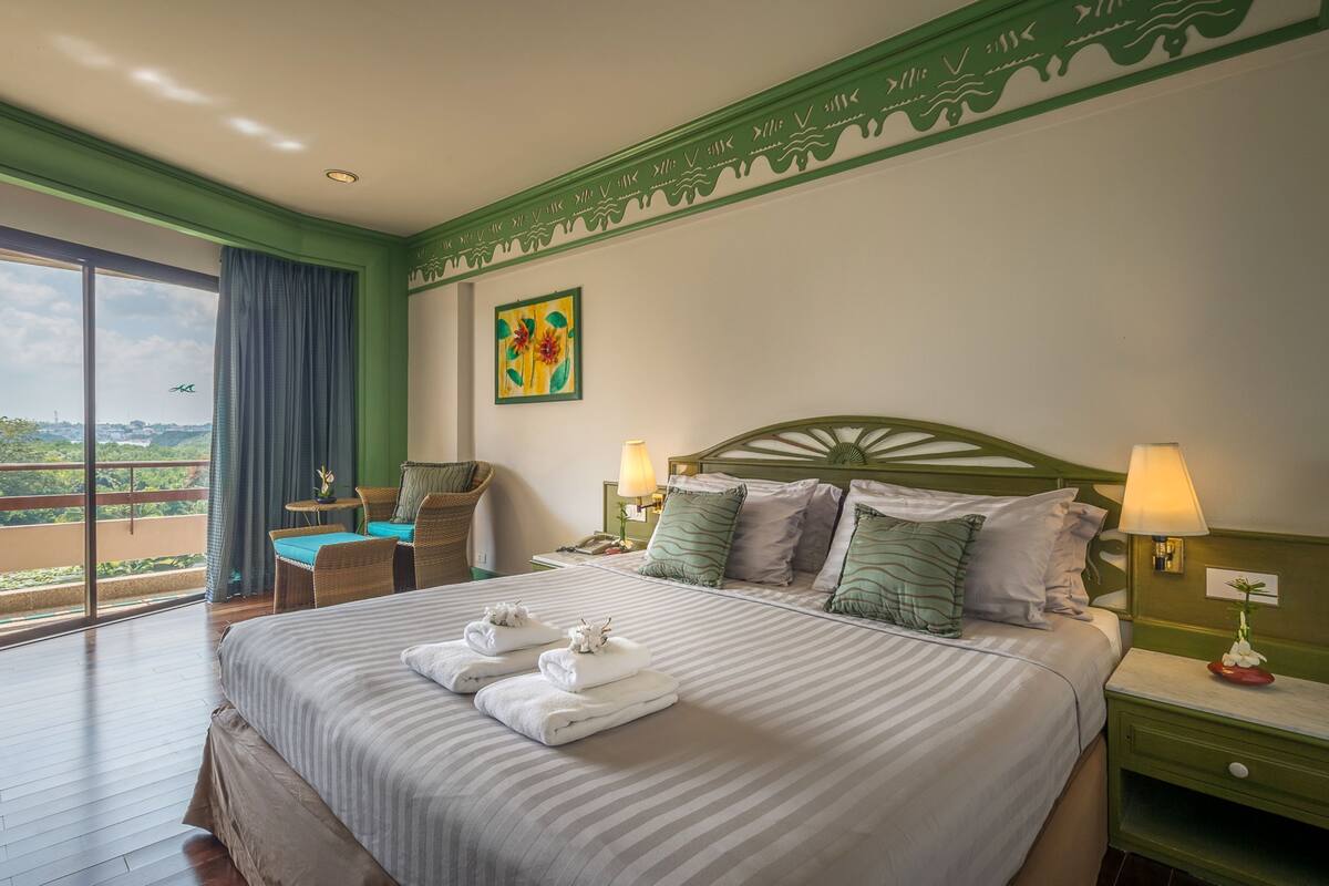 A hotel room with a large bed, two bedside lamps, a green headboard, a window with a view, and a small table with a chair.
