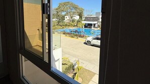 Une vue depuis une fenêtre d’un immeuble, avec une piscine, une voiture blanche garée à l’extérieur et un ciel bleu dégagé.