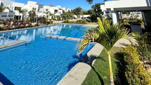 Une piscine entourée de bâtiments modernes, sous un ciel bleu azur, et bordée d’une végétation luxuriante.