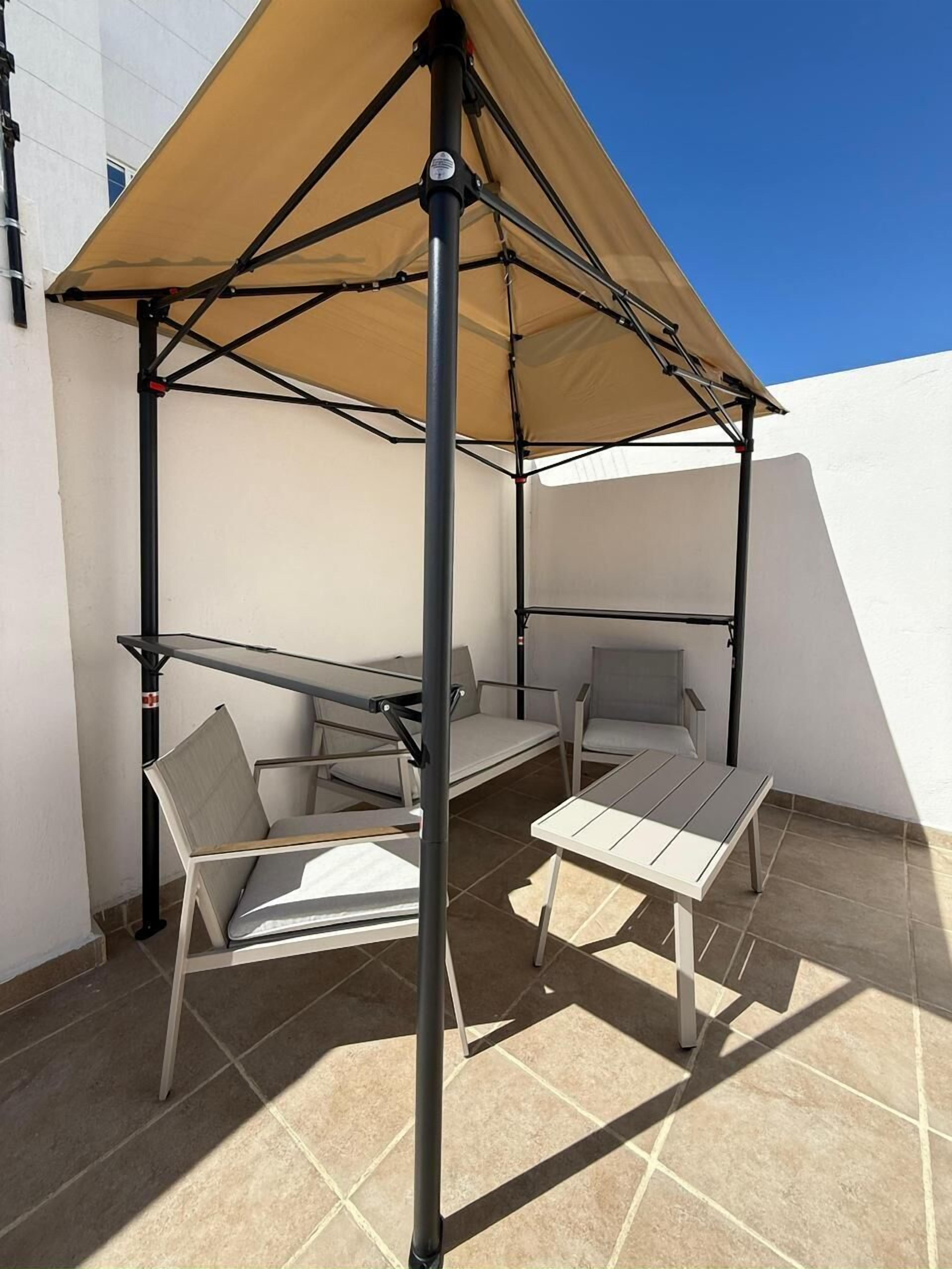 Un espace patio avec une table et deux chaises sous une marquise, un sol carrelé et un mur blanc.