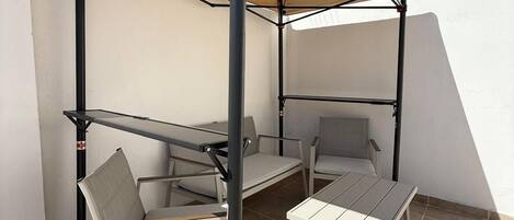 Un espace patio avec une table et deux chaises sous une marquise, un sol carrelé et un mur blanc.