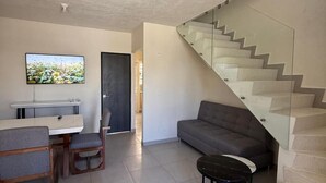 Un salon moderne avec un canapé gris, une table à manger avec des chaises, un téléviseur fixé au mur et un escalier en verre menant à un autre niveau.