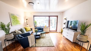 A modern living room with a sofa, coffee table, and TV. There are plants, a radiator, and a large window. - Beautiful house with conservatory and garden and large bathrooms (Brunsbüttel)