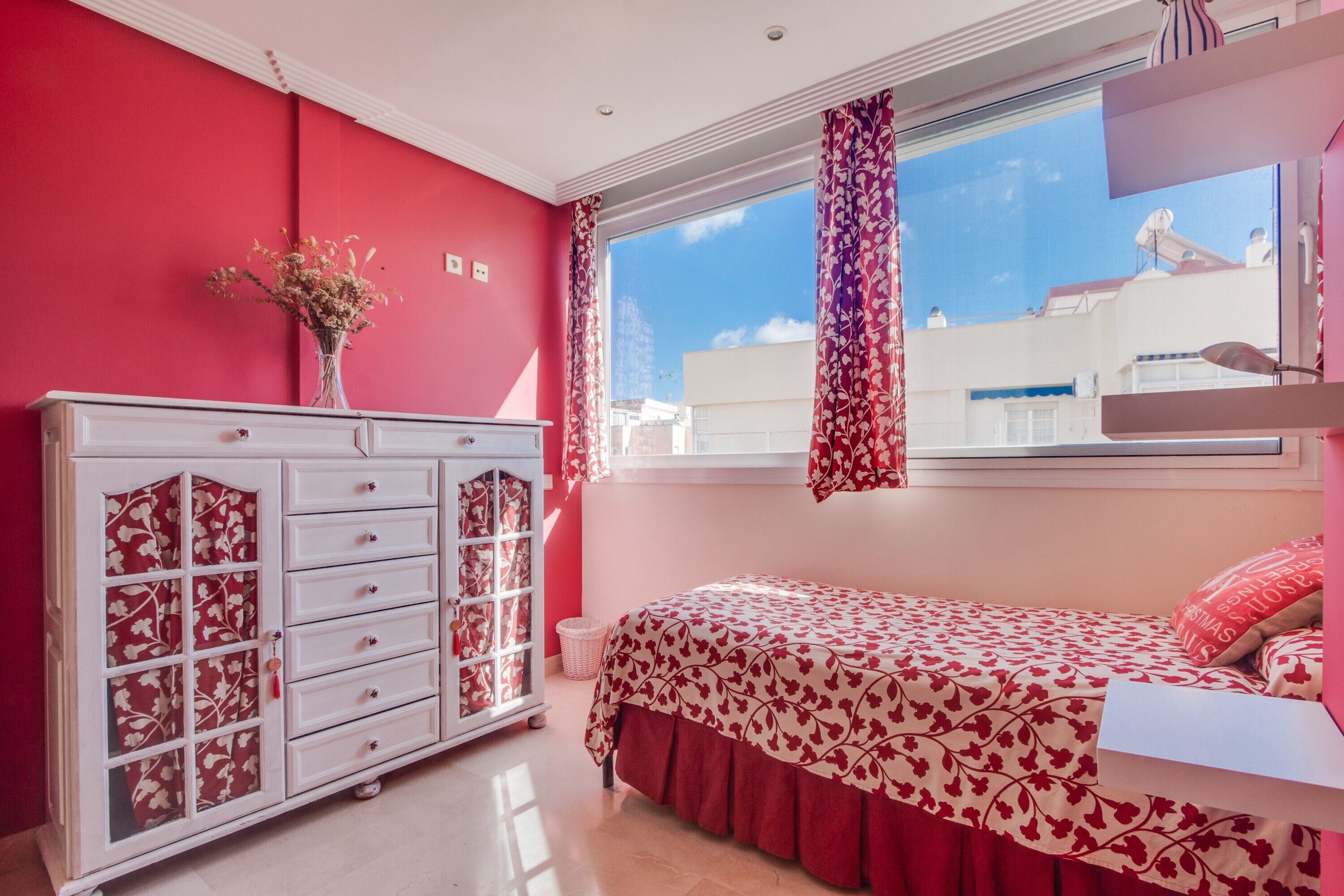 A bedroom with a bed, a dresser, a window with curtains, and a vase with flowers.