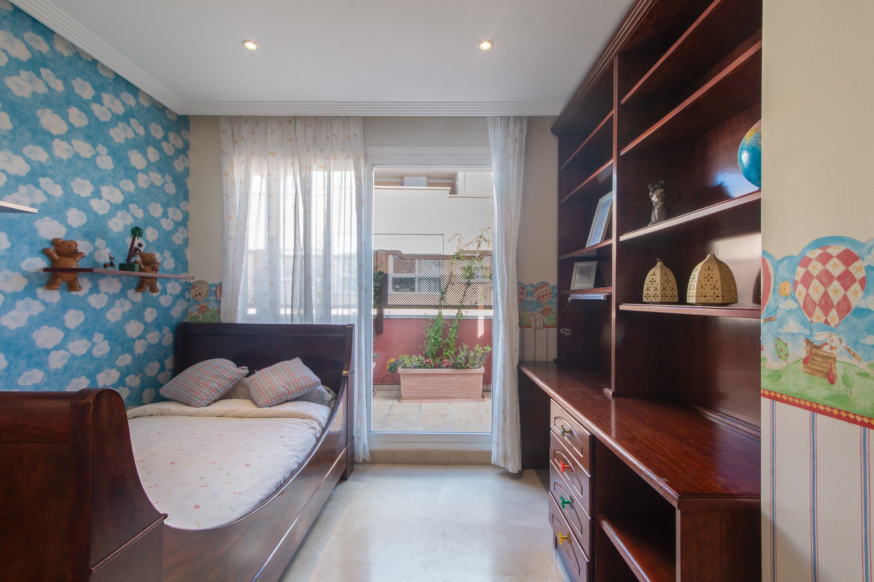 Une chambre à coucher comprenant un lit, une armoire, une étagère avec des objets décoratifs, une fenêtre avec des rideaux et une vue sur le balcon extérieur.