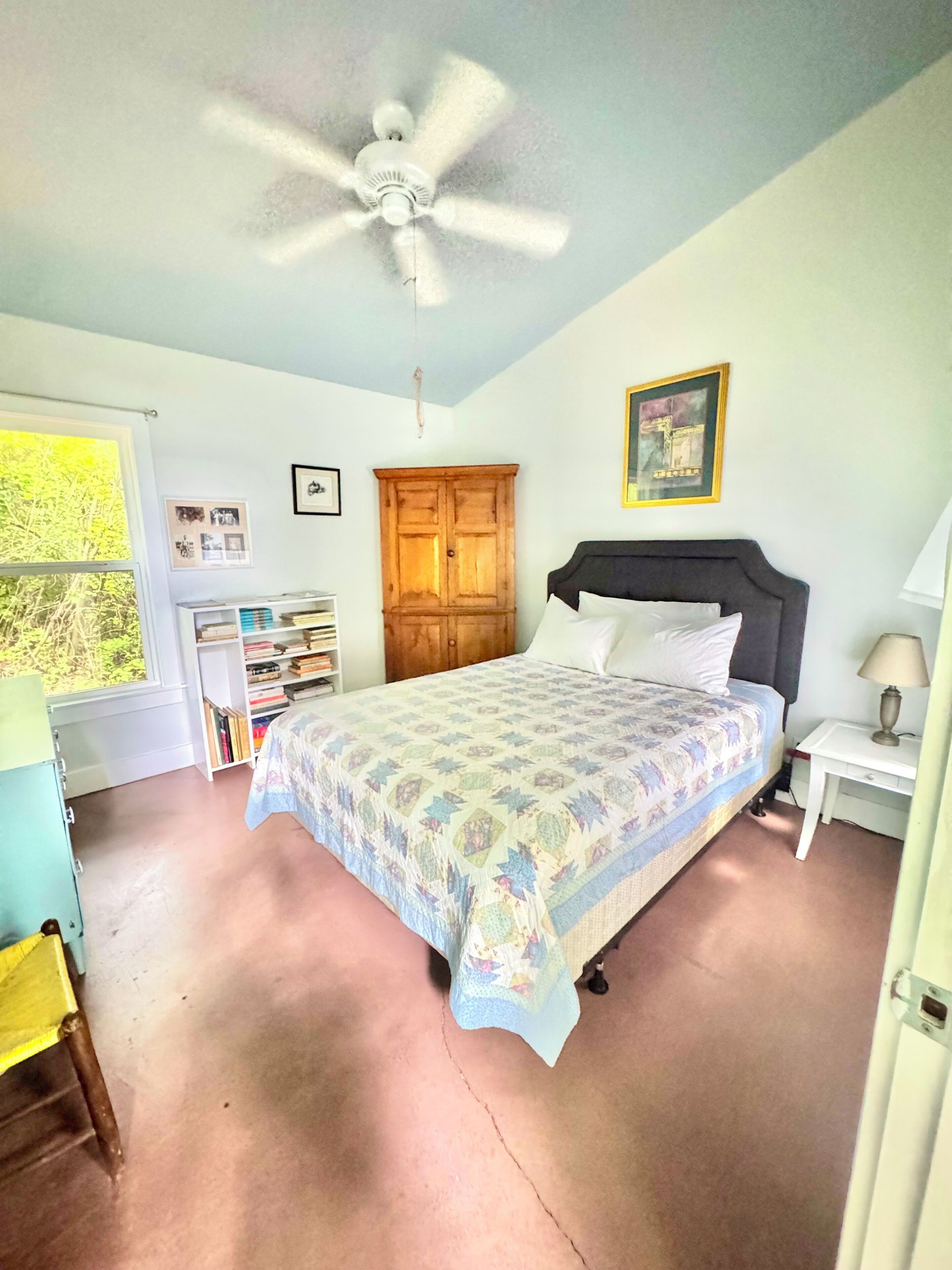 Ein Schlafzimmer mit einem Bett, einem Deckenventilator, einem Kleiderschrank, einem Schreibtisch und einem Fenster mit Blick auf Grünflächen.