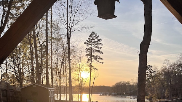 A hot tub with a rubber duck, a wooden deck, a small shed, and a view of a lake with a sunset. - "High Rock Haven- Fire Pit, Welcome Gifts,  Games room, Hot Tub, 2 lounges, lake (Lexington)