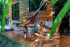 A hammock tied to a wooden rocking chair, set on a tiled patio with a wooden house in the background.
