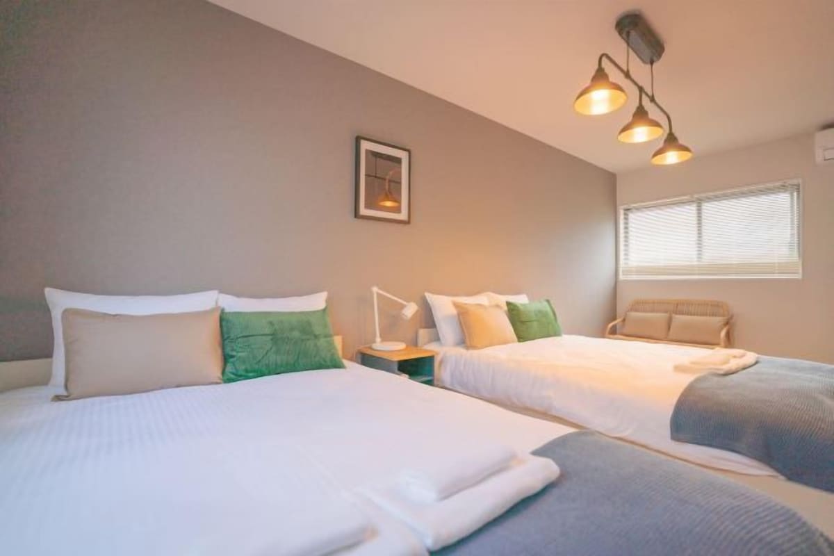 A hotel room with two beds, a bedside table with a lamp, and a window with blinds.