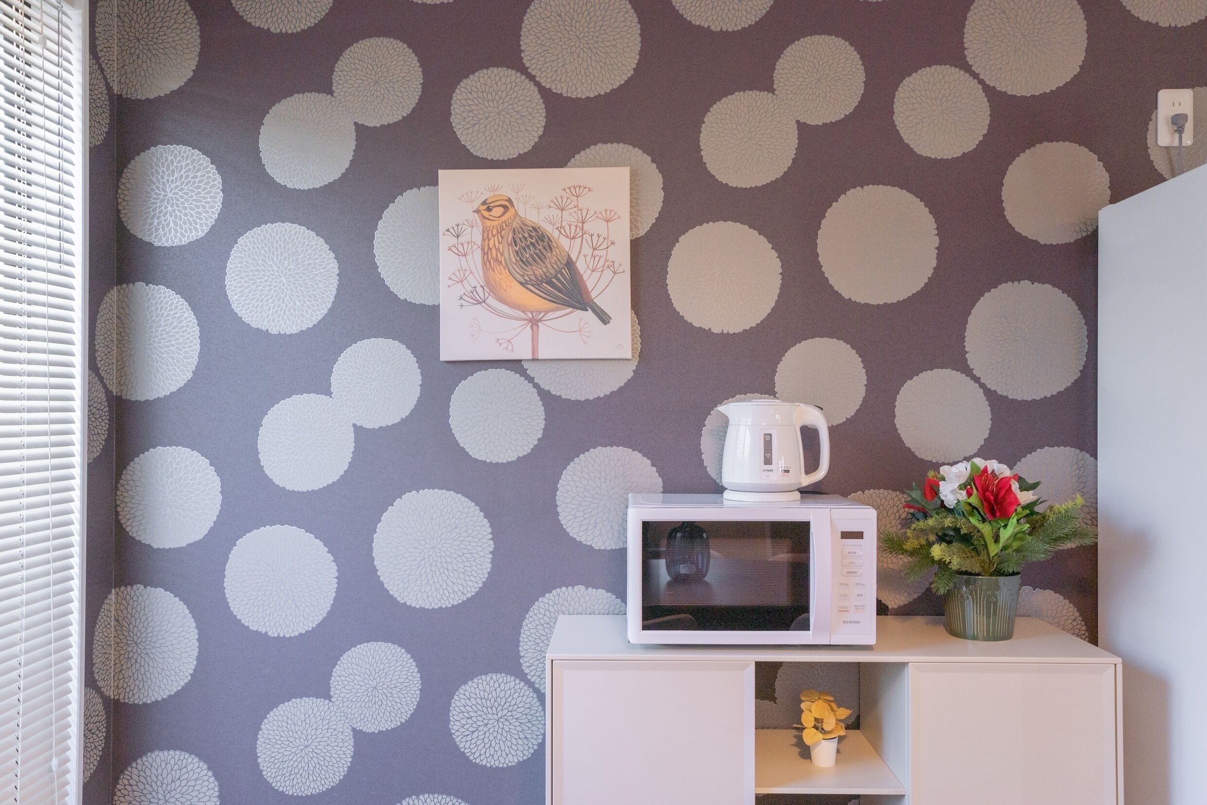 A room with a polka dot wallpaper, a white cabinet with a microwave and a vase of flowers.