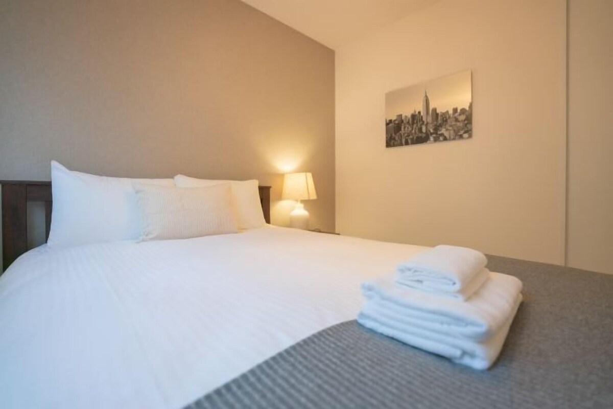A neatly made bed with white linens, a folded towel on the side, and a cityscape painting on the wall.
