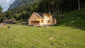 Una cabaña de madera con un porche y una mesa, rodeada por un campo cubierto de hierba y la silueta de una montaña boscosa al fondo.
