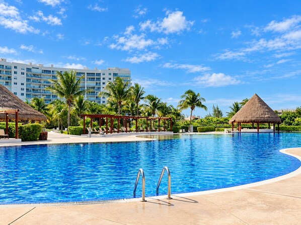 A swimming pool with a clear blue water, surrounded by lounge chairs and palm trees, with a hotel building in the background.