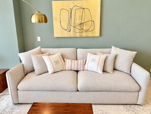 Ein helles Sofa mit vielen Kissen, eine goldene Pendelleuchte und ein großes abstraktes Kunstwerk an der Wand.