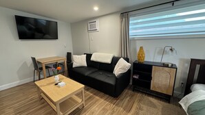 Ein modernes Hotelzimmer mit einer schwarzen Couch, einem Holztisch, einem Fernseher, einer Klimaanlage, einem Nachttisch mit Lampe und einem Fenster mit Jalousien.