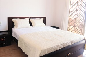 A neatly made bed with a white bedspread and two pillows, a wooden headboard, a nightstand with a drawer, and a window with a geometric pattern. - Hotel - Casa del Mar Malin en Santa Elena, Ballenita, al frente al Mar (Ballenita)