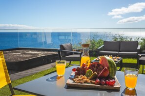 Un tavolo con un piatto di frutta, due bicchieri di succo d'arancia e una bottiglia di succo, su un balcone con vista sull'oceano.