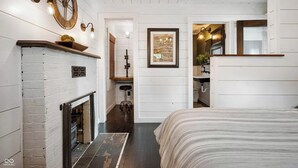A bedroom with a fireplace, a bed, a bathroom, and a hallway with a clock and wall art.
