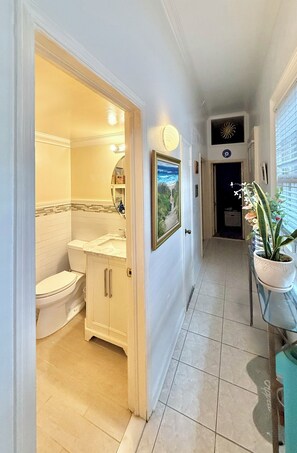 Un couloir mène à une salle de bain équipée de toilettes, d’un lavabo et de placards. Une plante en pot est posée sur une table, et un tableau encadré orne le mur.