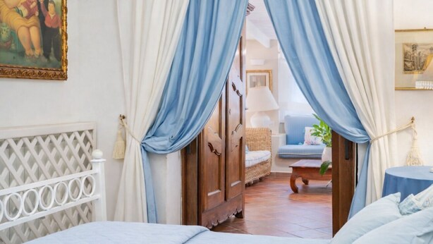 A bedroom with a bed, a wooden wardrobe, and a view into another room with a sofa and a lamp.
