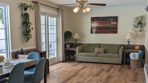 A cozy living room with a wooden floor, a green sofa, a dining table with chairs, and a ceiling fan.