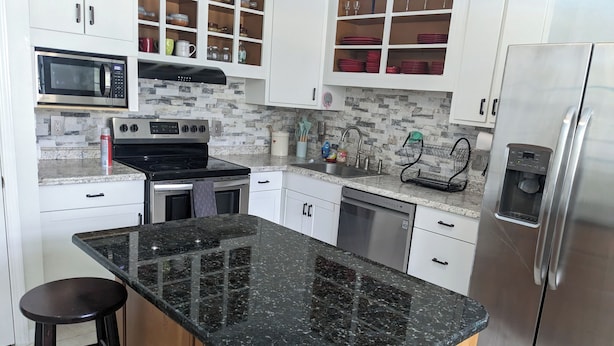 A modern kitchen with white cabinets, a black granite countertop, stainless steel appliances, and a wooden stool.