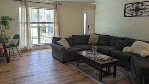 A living room with a dark grey sofa, a wooden coffee table, and a large window with curtains.