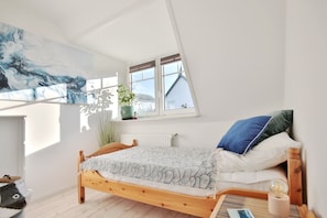 Quarto com teto inclinado, cama de madeira, janela com vista para um edifício e uma grande pintura abstrata na parede.