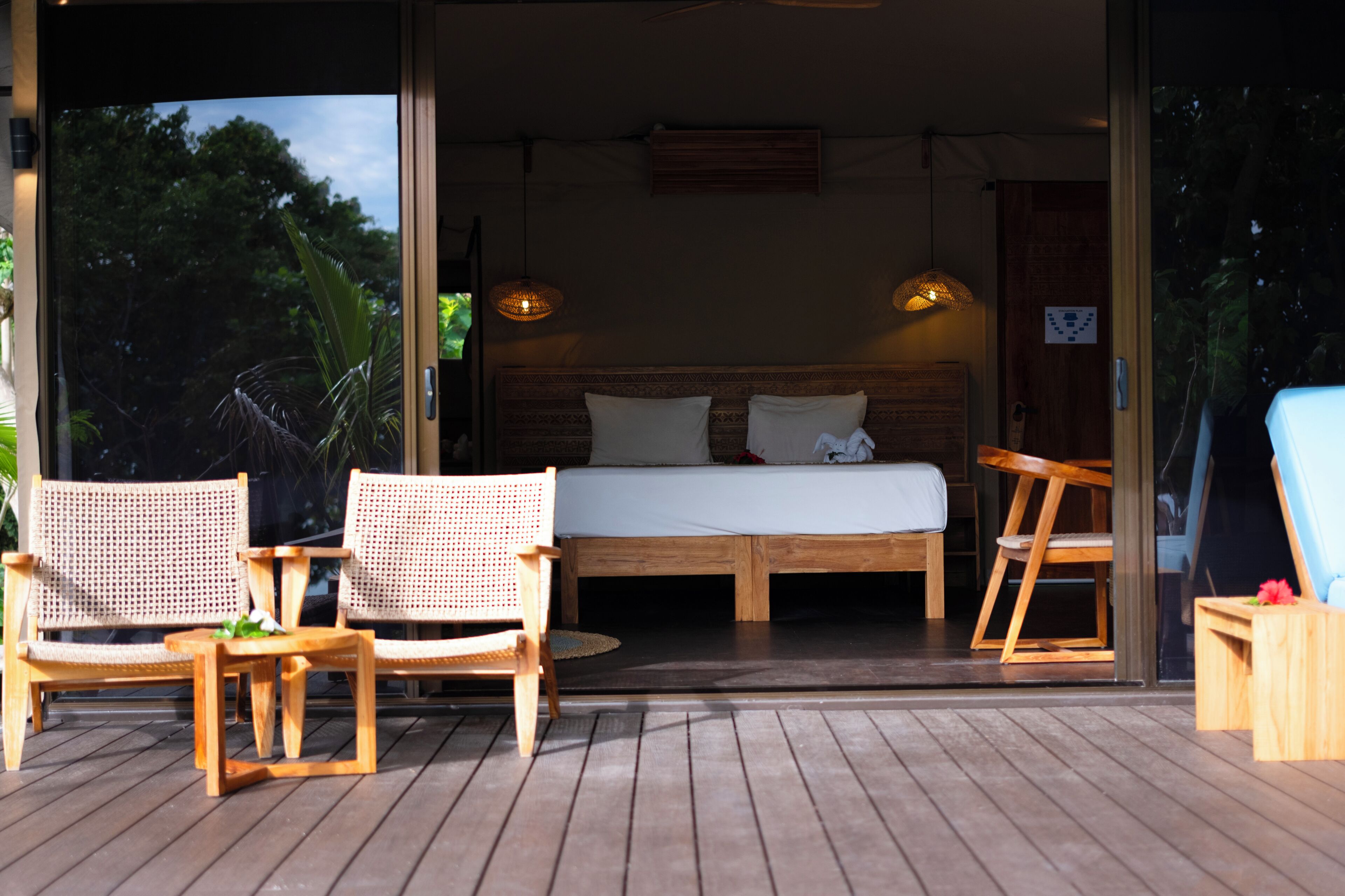 An open-air bedroom with a bed, two wicker chairs, a wooden table, and a view of trees.