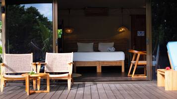 An open-air bedroom with a bed, two wicker chairs, a wooden table, and a view of trees.