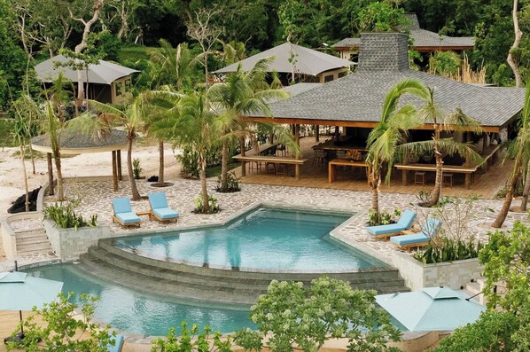 Zona de piscina con sillones reclinables, un espacio de descanso cubierto y un edificio con tejado de tejas.