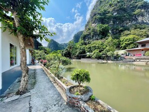 A riverside hotel with a paved walkway, a small garden, and a view of the surrounding mountains. - Trang An Pristine View Homestay (Hoa Lu)