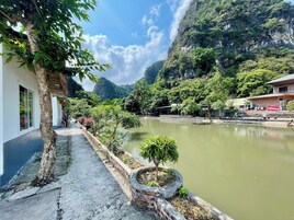 Um hotel à beira do rio, com um passeio pavimentado, um pequeno jardim e vista para as montanhas ao redor.