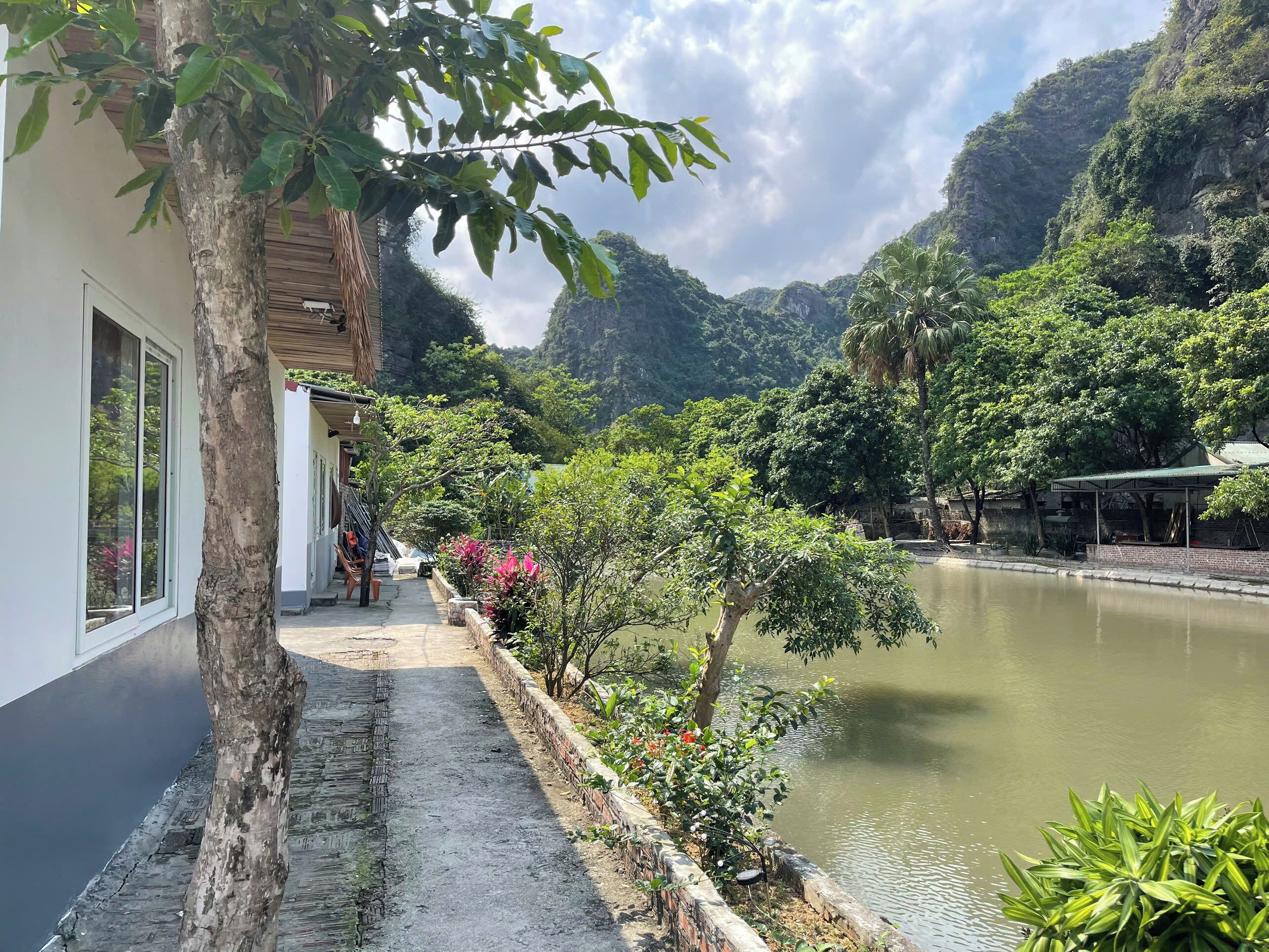 A riverside walkway with a building on one side, a bridge over the water, and lush greenery in the background.