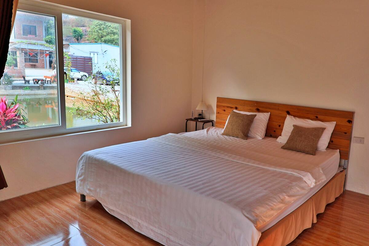 Una cama bien hecha con cabecera de madera, una mesita de noche con lámpara y una ventana que ofrece vistas a un edificio y vegetación.
